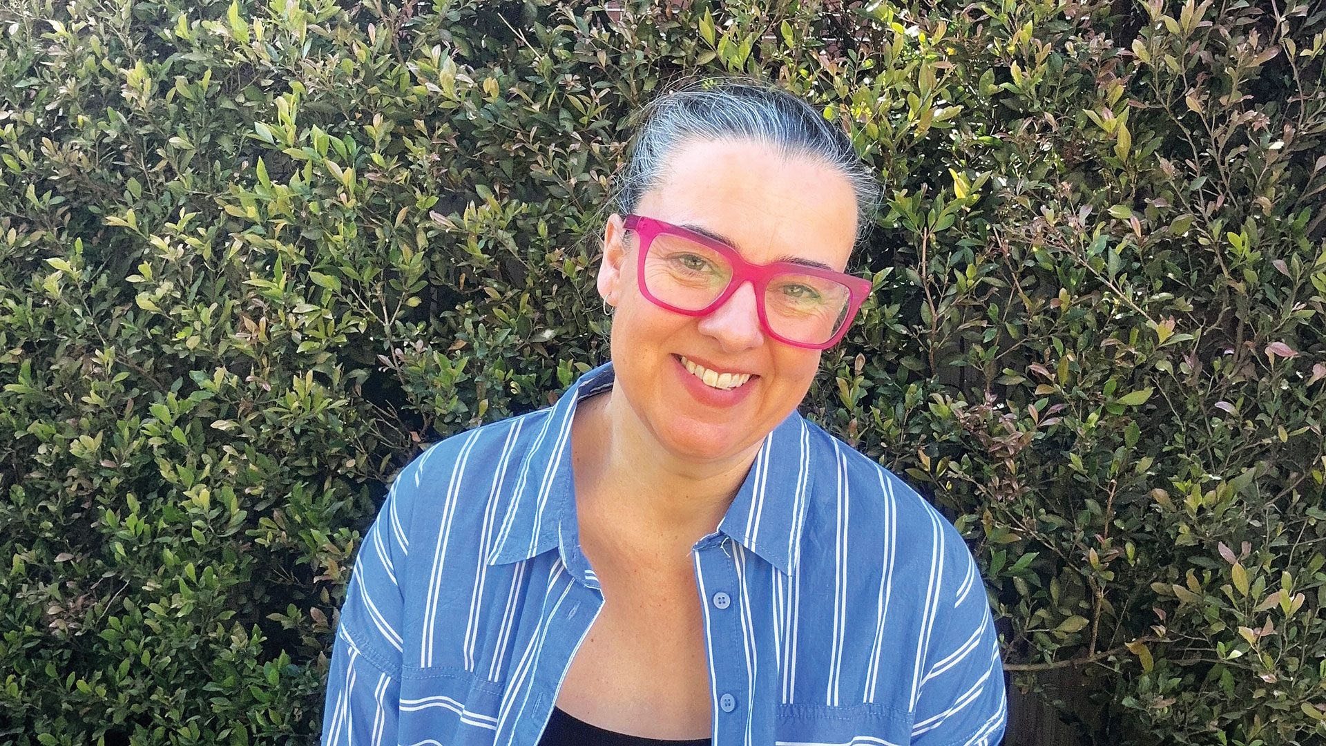 Person with pink glasses smiling in front of a green hedge, wearing a blue and white striped shirt.