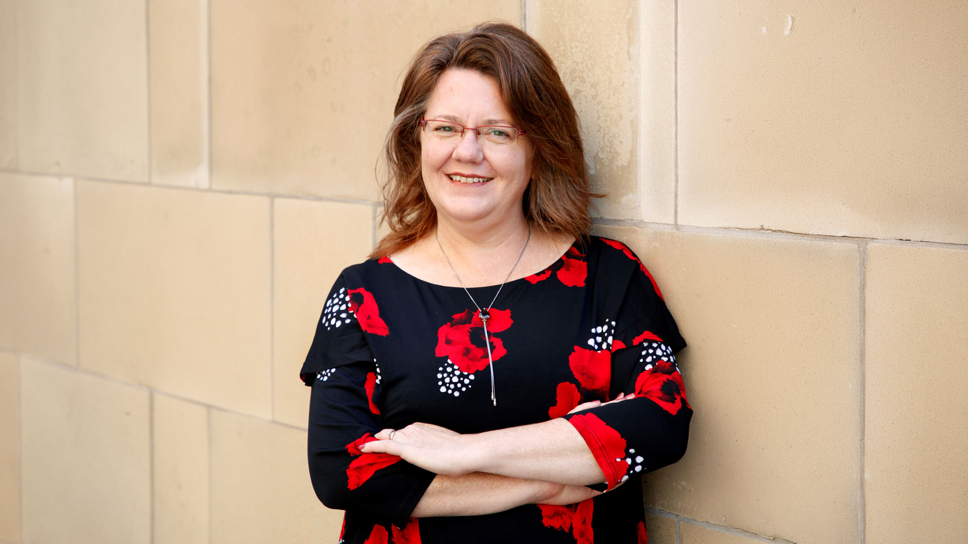 Woman with red glasses smiling and leaning against a beige wall, wearing a black dress with red floral print
