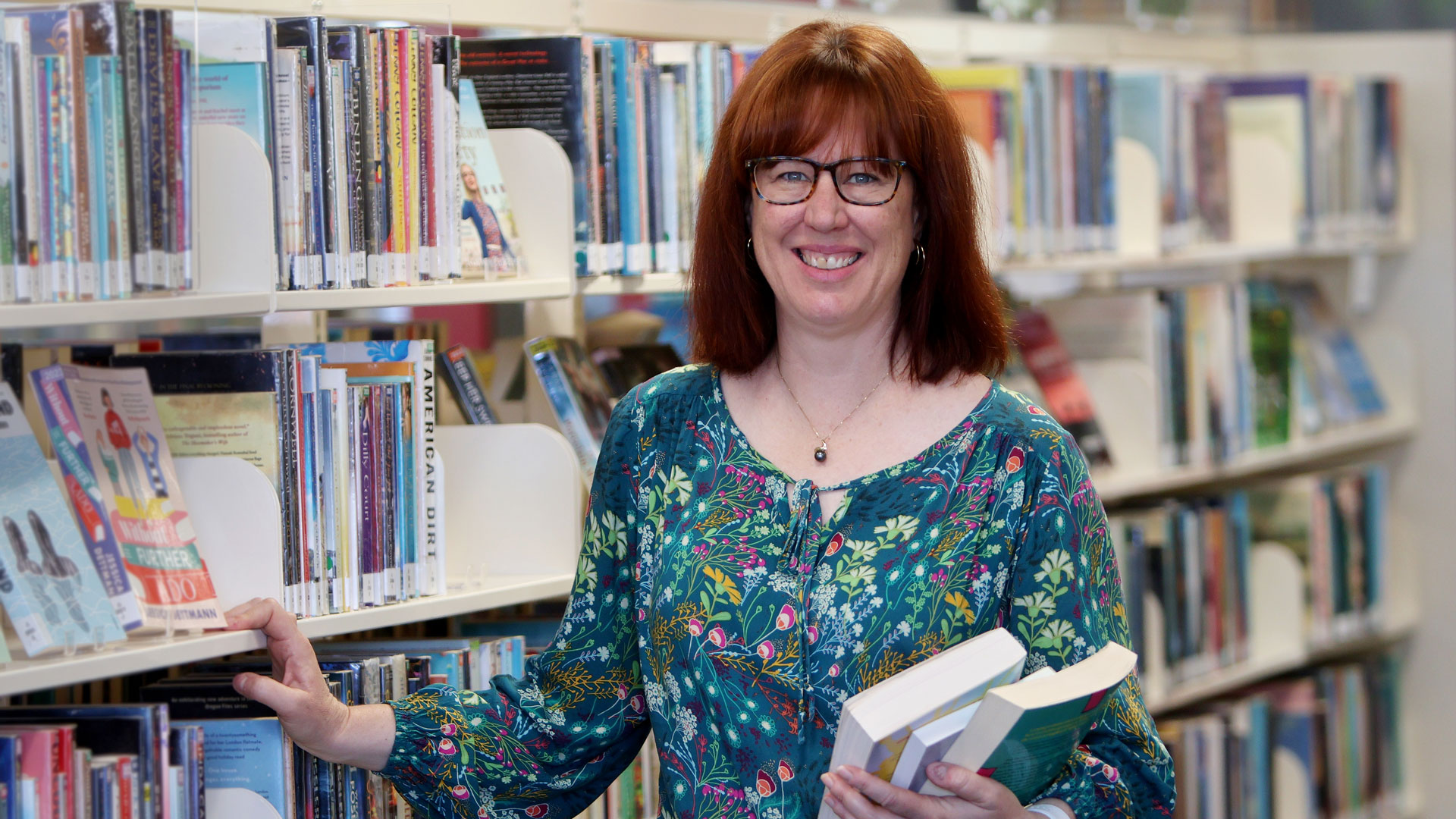 Melissa Whitrow holding three books, standing in front of a library shelf