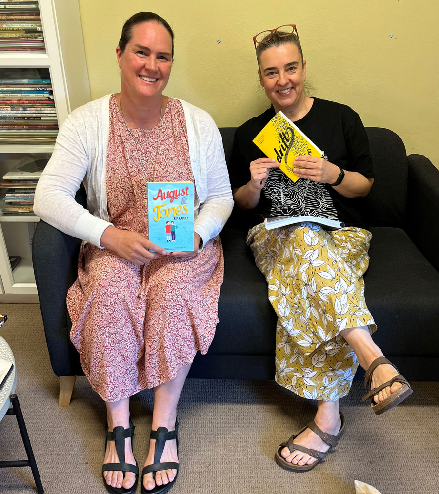 Two women in a library setting sitting on a couch and holding books, smiling at the camera.