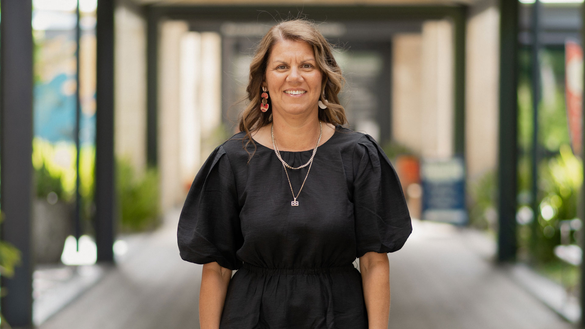 Woman smiling outdoors, wearing a black dress and standing on a walkway with blurred buildings in the background