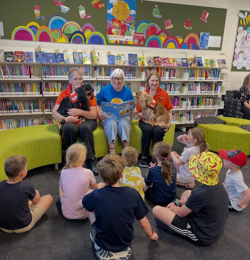 Children listen to a story being read at a public library during a Big Summer Read event, with two therapy dogs and their handlers seated alongside the reader.