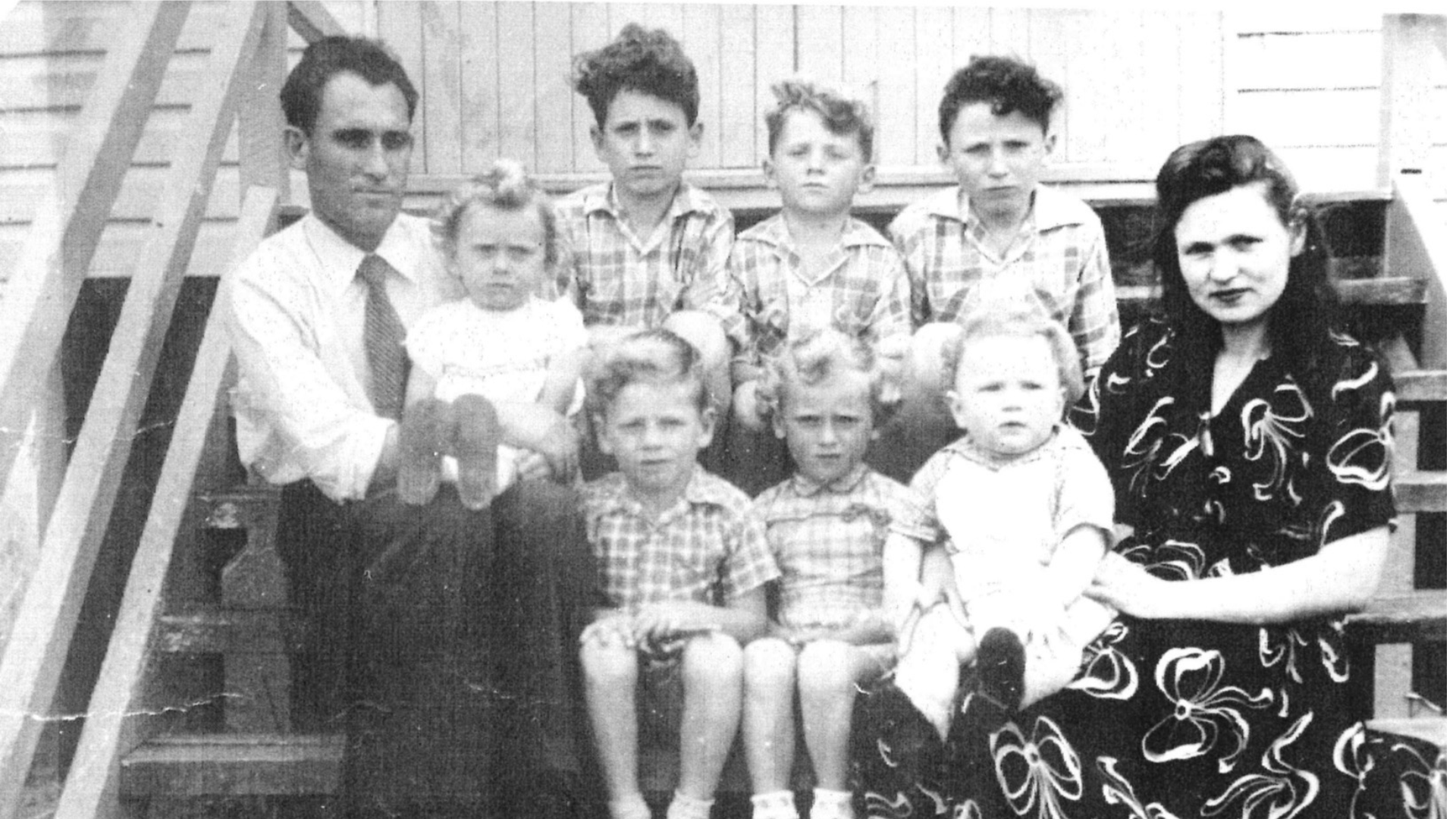 Black-and-white historical family photograph showing two adults and seven children seated on the steps of a timber house. The father sits on the left holding a young child, while the mother sits on the right with a toddler on her lap. Five other children, ranging from toddlers to teenagers, are arranged between and behind them on the steps. Most of the children wear matching or similar patterned clothing.