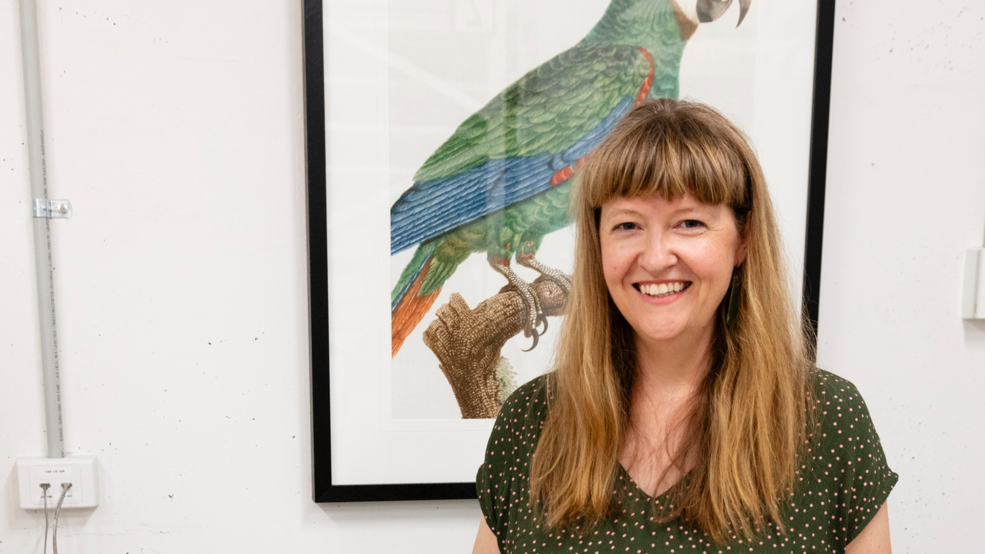 Person smiling in front of a framed illustration of a green and blue parrot.
