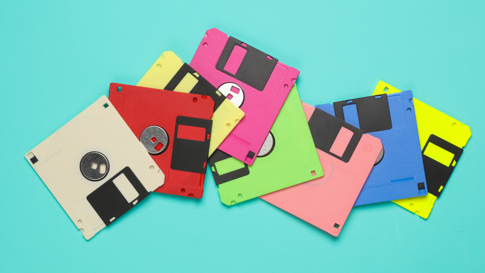 A collection of brightly coloured floppy disks in various sizes is scattered across a turquoise background. The image evokes early digital storage technologies and the evolution of data preservation from physical media to modern digital formats.
