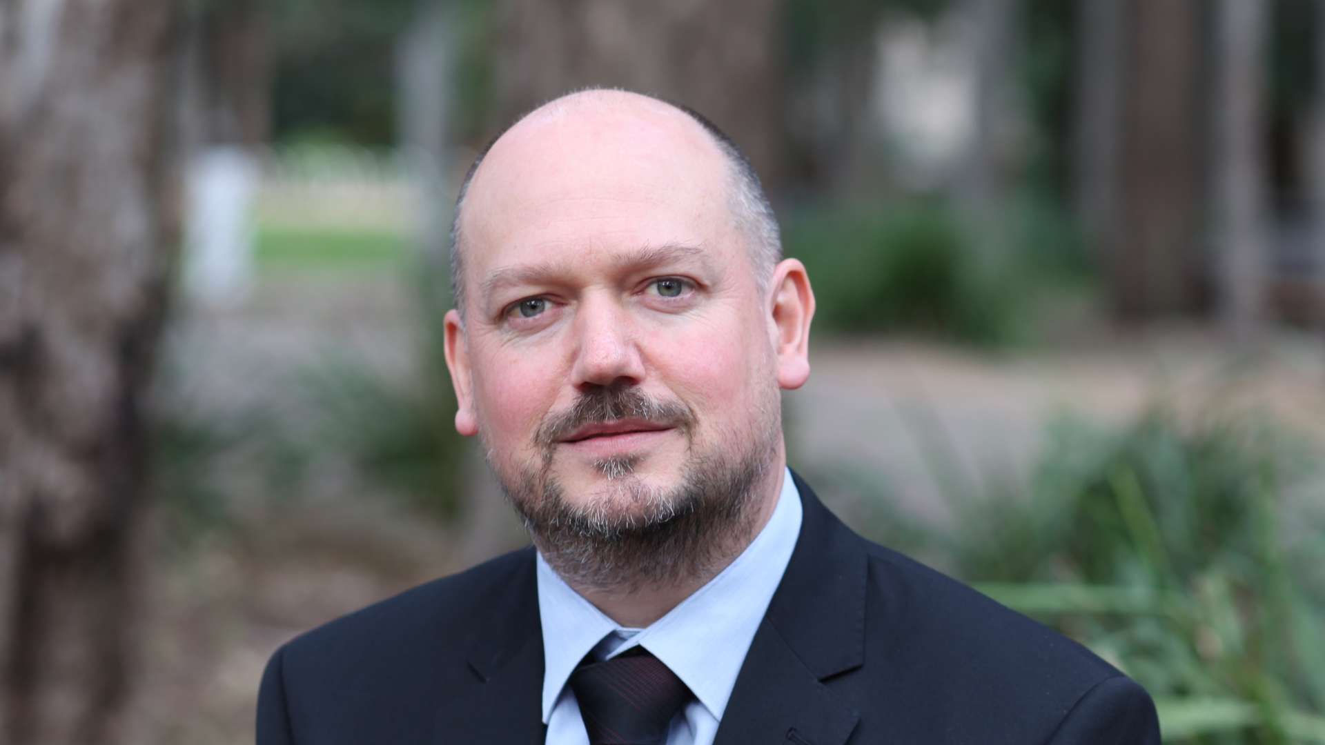 Head-and-shoulders portrait of a man in a dark suit, light blue shirt and tie, looking directly at the camera. He is photographed outdoors with trees and greenery softly blurred in the background.