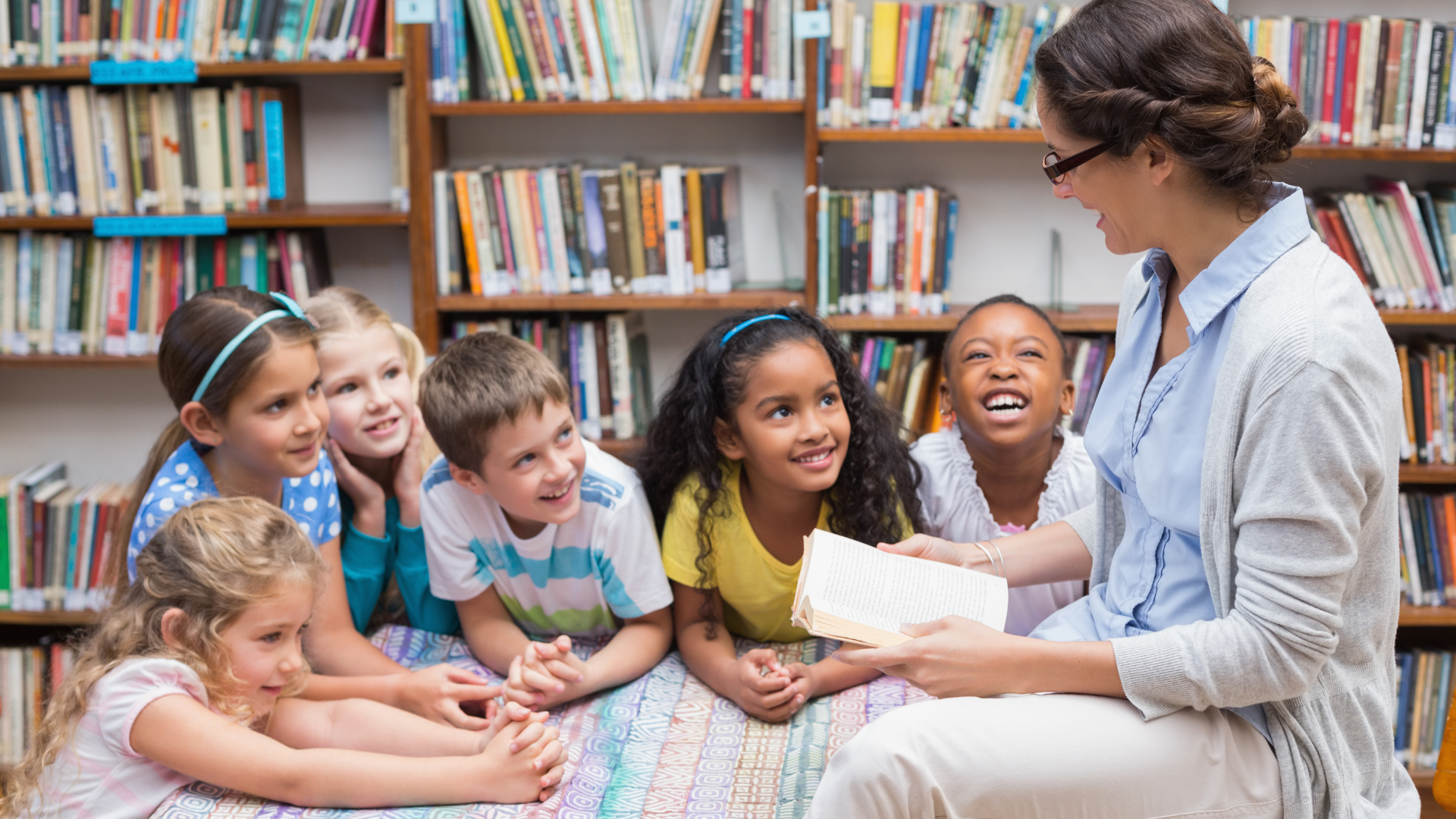 A librarian sits on the floor of a library reading aloud from a book to a group of six young children. The children, from diverse backgrounds, are gathered closely around her, smiling, laughing and listening attentively.