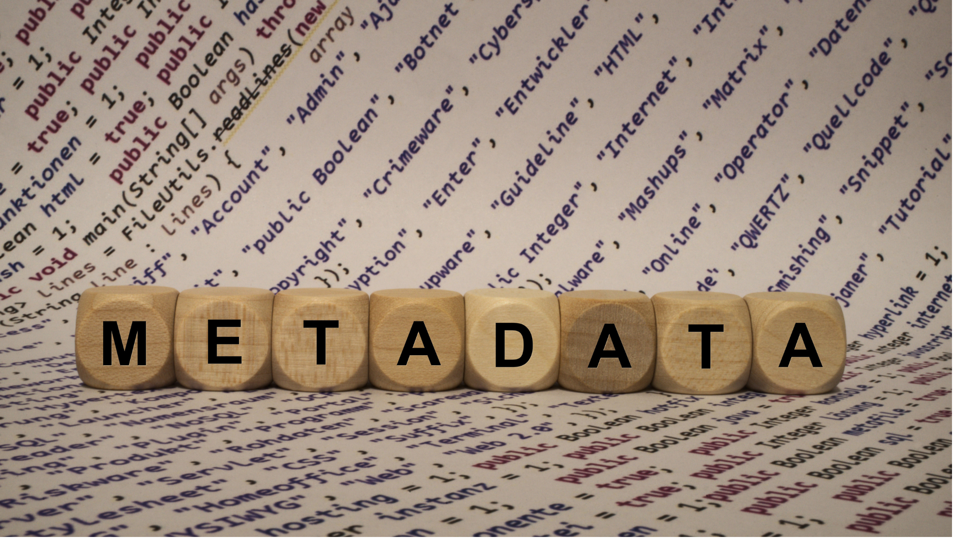 Wooden letter blocks spelling “METADATA” arranged in a row on top of a printed page of computer code and technical keywords. The image symbolises the role of metadata in organising, describing and managing digital information.