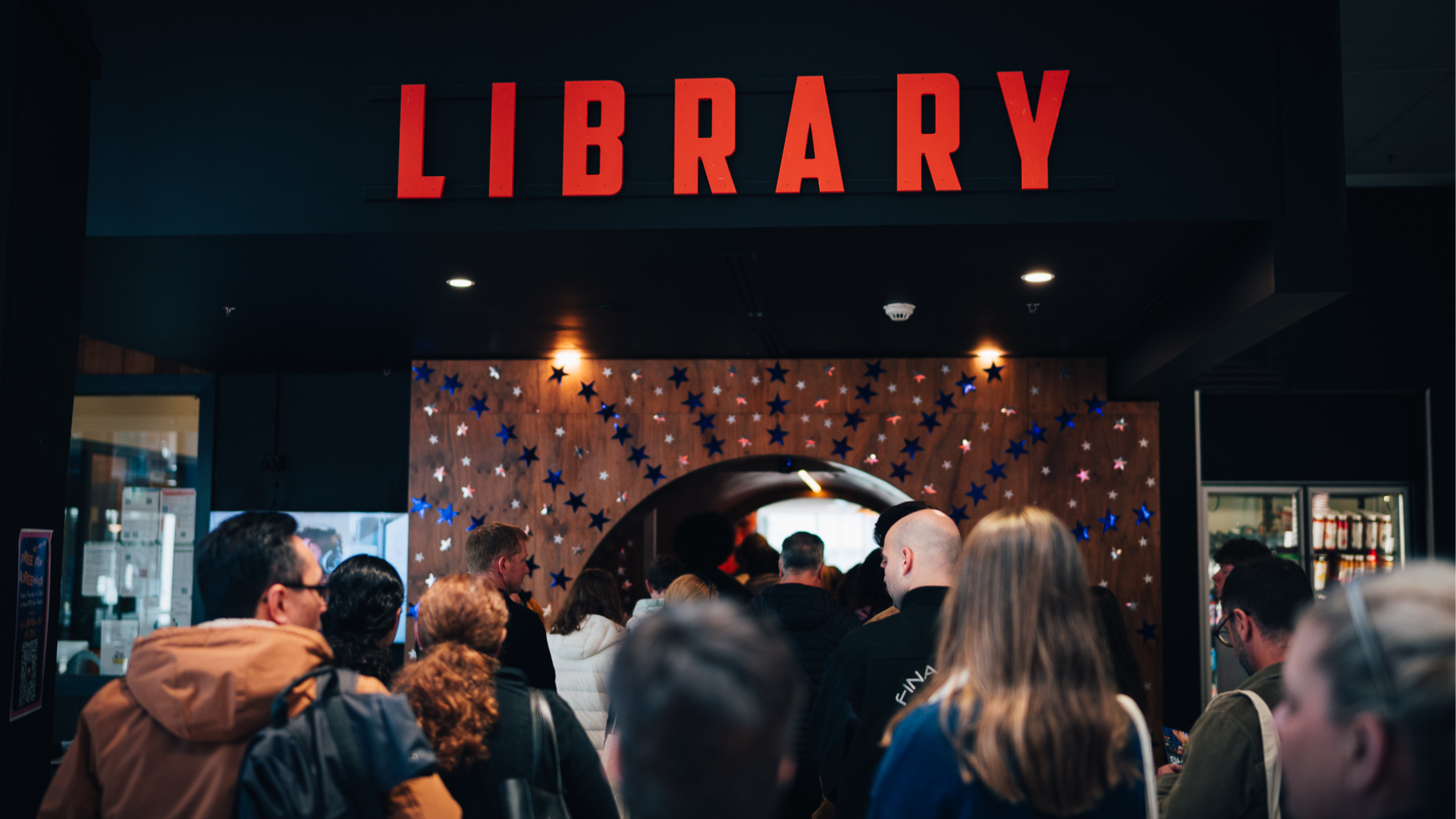 A crowd of people enters a library through a wide entrance beneath a large illuminated red "LIBRARY" sign. The doorway is decorated with blue and silver star-shaped decorations, creating a welcoming atmosphere as visitors make their way inside.