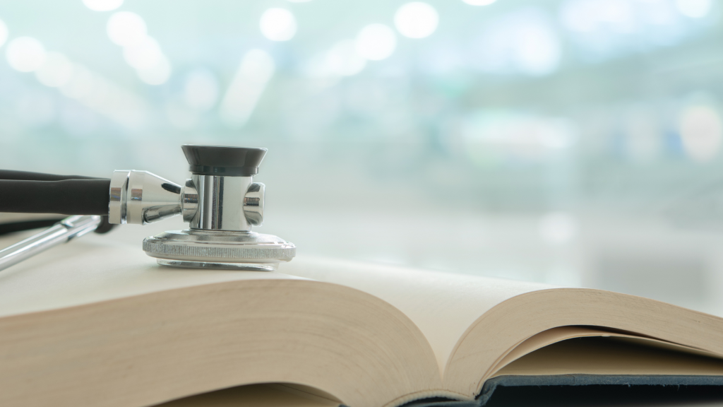 A silver stethoscope rests across the open pages of a thick book, symbolising the connection between healthcare and knowledge. The softly blurred background features cool blue and white tones with circular bokeh lights, creating a calm, clinical atmosphere.