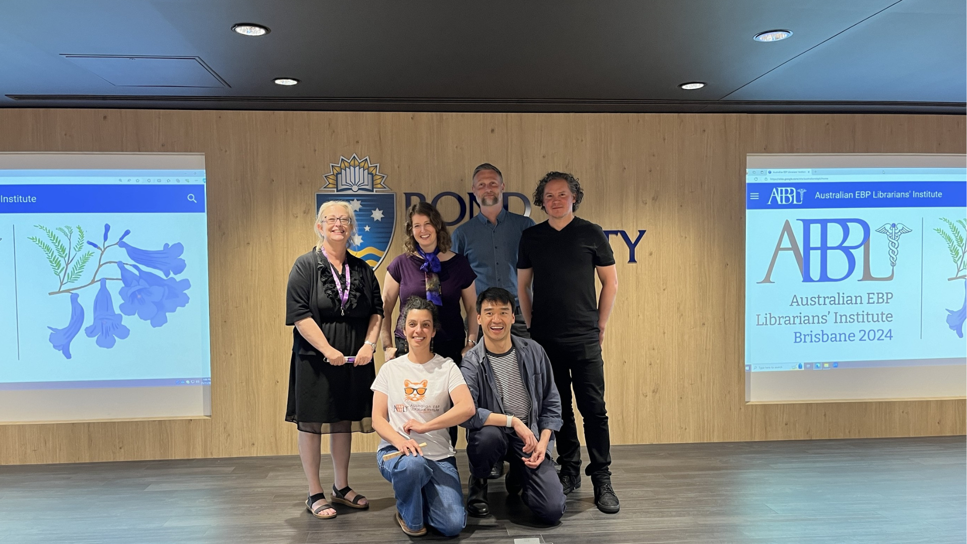 AEBPLI Hero Image Six people pose together at the Australian EBP Librarians’ Institute Brisbane 2024, with event branding displayed on screens on either side.