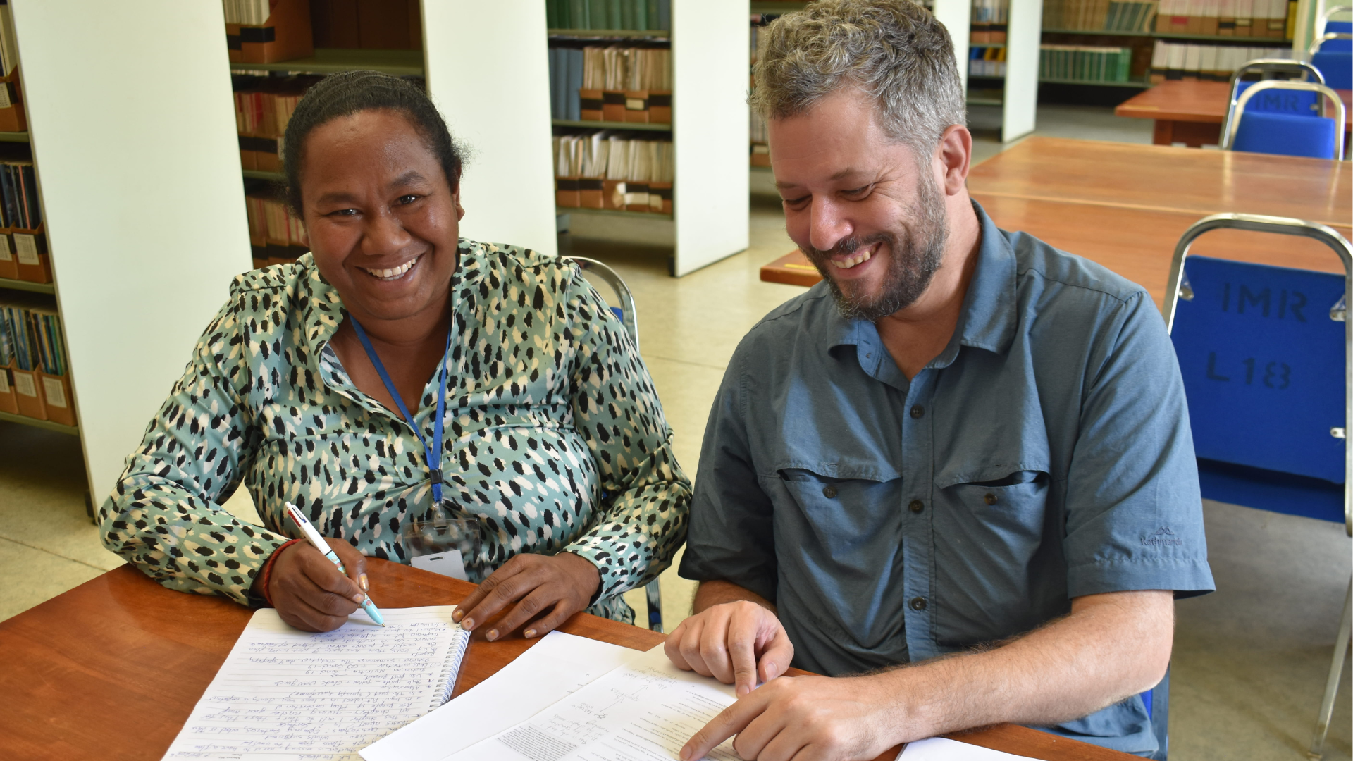 Two people smile while reviewing handwritten notes and documents together at a table in a library.