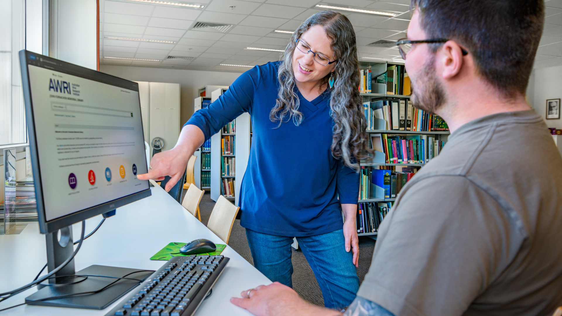 A woman points to information on a computer screen while assisting a seated man in a library or information centre, with bookshelves in the background.