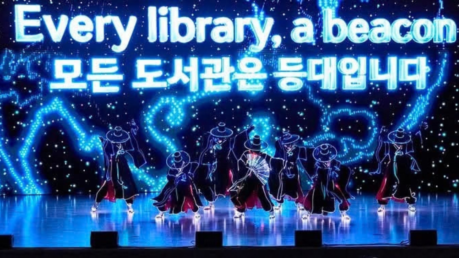 Performers in dark costumes and traditional hats dance on a brightly lit stage, beneath the words “Every library, a beacon” in English and Korean.
