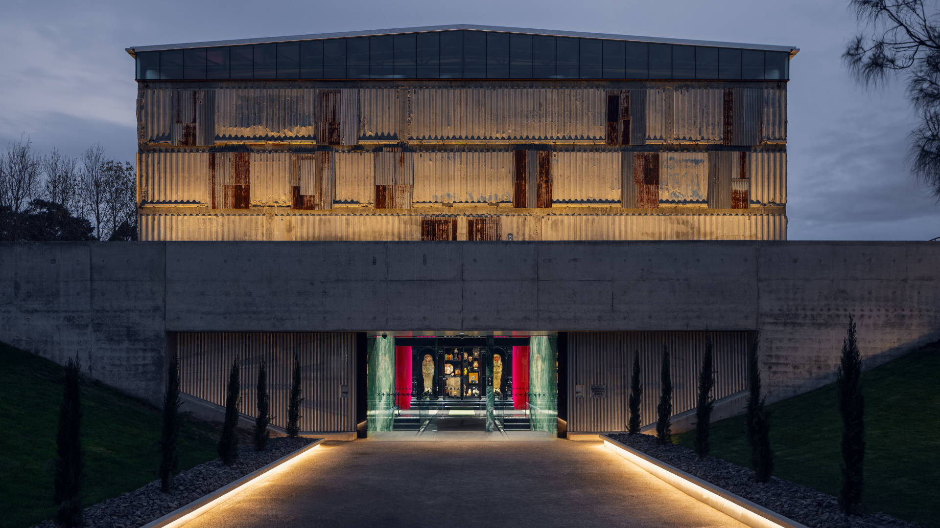 Exterior of MONA at dusk, showing the illuminated corrugated metal building above a concrete entrance, with a lit pathway leading inside.